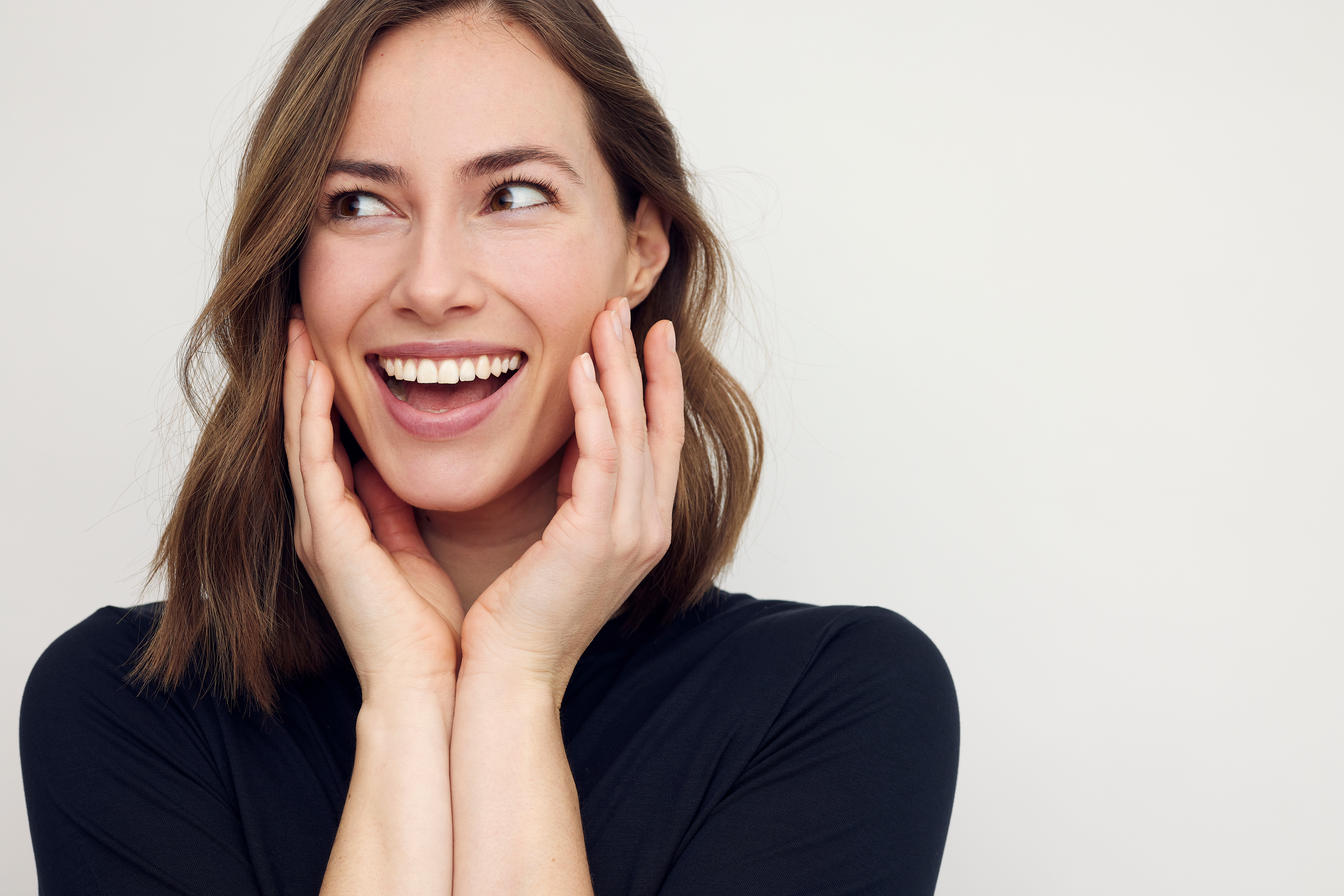 Portrait of a beautiful happy woman smiling and thinking, with hands on her cheeks like she’s thinking and making a surprised expression. Female in black with big smile isolated on white background. woman with an excited expression